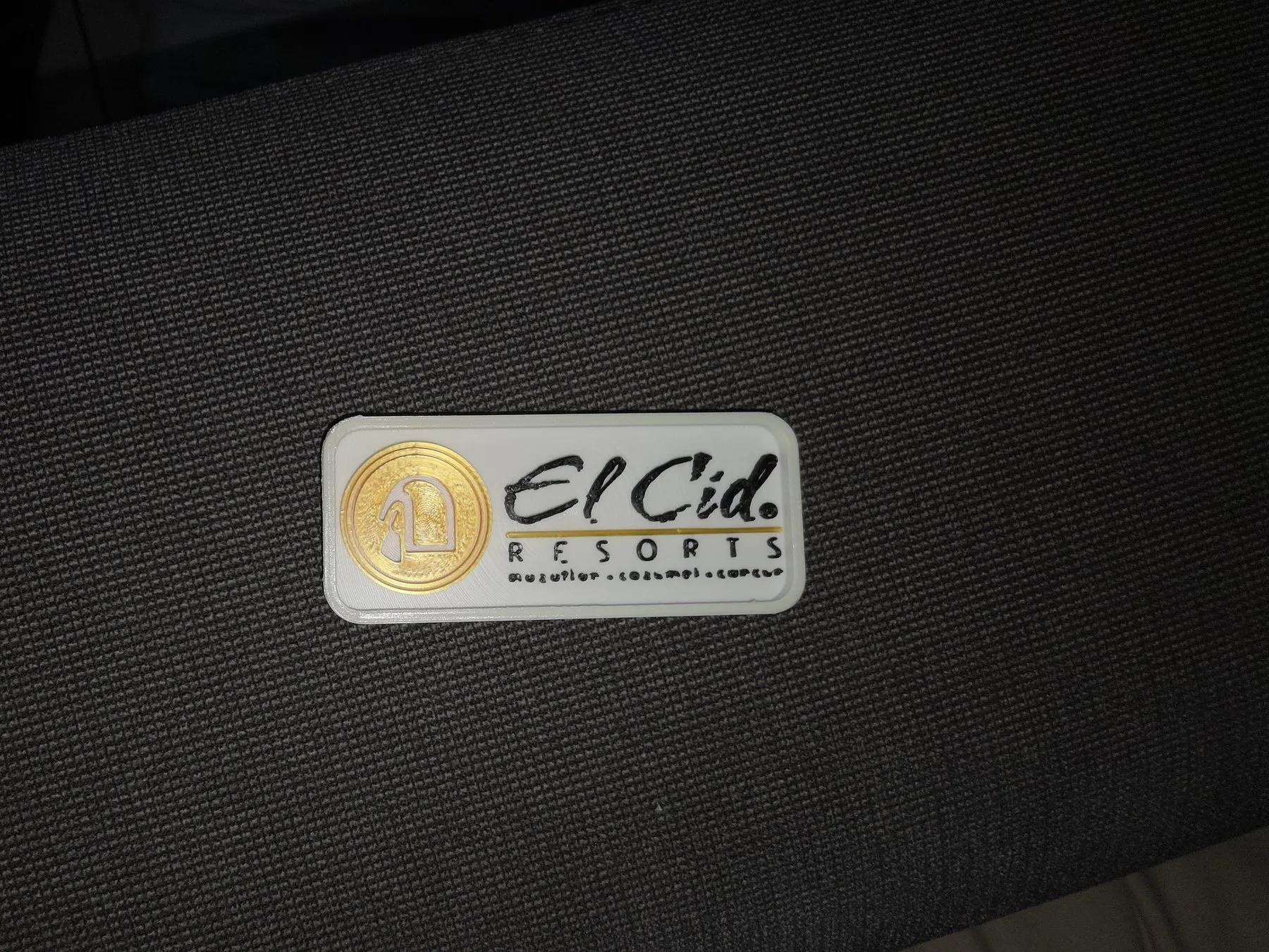 Bag tag blanco de El Cid Resorts con el logotipo en negro y una moneda dorada, sobre tela oscura